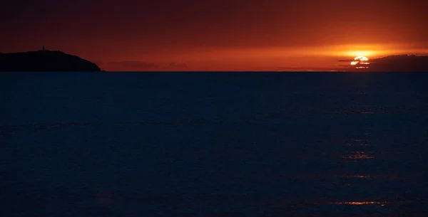 El faro en la montaña al atardecer, el cielo anaranjado, el mar tranquilo, el camino del sol — Foto de Stock