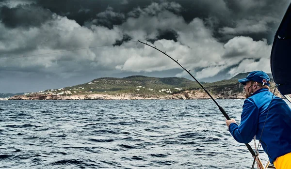 El pescador pesca desde el barco, está vestido con chaqueta azul y gorra, el paisaje del mar baleárico y las montañas improbables, el agua azul, el cielo de tormenta — Foto de Stock