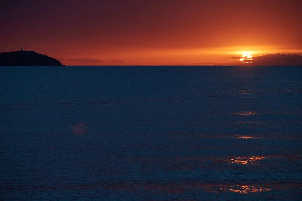 A világítótorony a hegyen naplementekor, a narancssárga ég, a csendes tenger, a napsütés útja. — Stock Fotó
