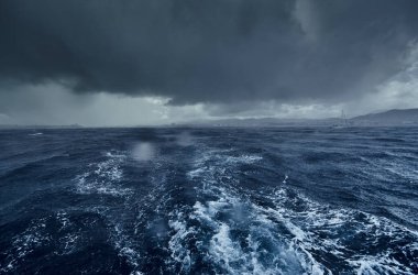 Fırtınalı deniz manzarası ve yelkenliden dağlar, bottan sonra köpüklü yol, teknenin altından fışkıran su, yağmurlu hava, dramatik gökyüzü.