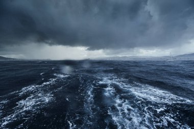 Fırtınalı deniz manzarası ve yelkenliden dağlar, bottan sonra köpüklü yol, teknenin altından fışkıran su, yağmurlu hava, dramatik gökyüzü.
