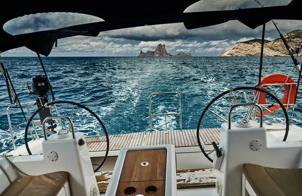 The view of the sea and mountains from the sailboat, edge of a board of the boat, oat stern, steering wheel, without people, slings and ropes, splashes from under the boat, sunny weather, dramatic sky