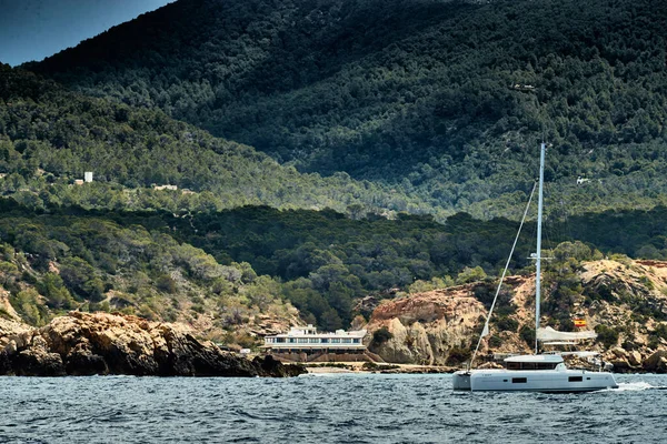 The Landscape of the balearic sea and improbable mountains, azure water, the storm sky, lonely buildings at tops of mountains, sail boat is on background — Stock Photo, Image