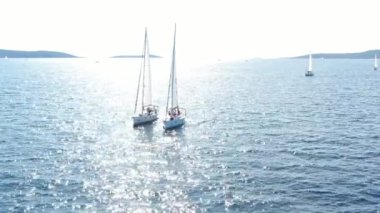 Birbirine yakın iki yelkenli yatı, gençlerin yatlarda eğlenmesi, gençlerin botların arasında asılı kalması, Adriyatik Denizi, Hırvatistan, arka planda adalar, suda güneş yansımaları, sükunet.