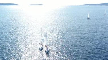 Birbirine yakın iki yelkenli yatı, gençlerin yatlarda eğlenmesi, gençlerin botların arasında asılı kalması, Adriyatik Denizi, Hırvatistan, arka planda adalar, suda güneş yansımaları, sükunet.