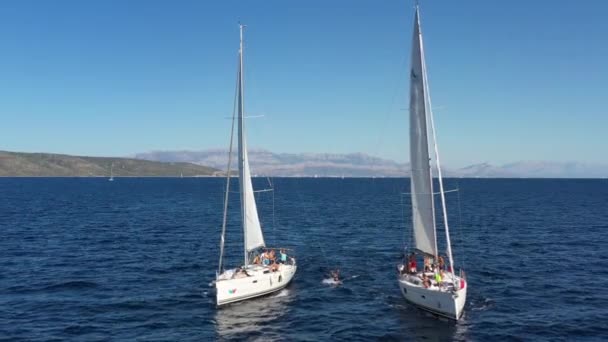 Хорватія, Адріатичне море, 18 вересня 2019 року: дві вітрильні яхти близько одна до одної, молодь веселиться на яхтах, молоді люди висять на мотузці між човнами, островами на задньому плані, сонце відбивається на воді. — стокове відео