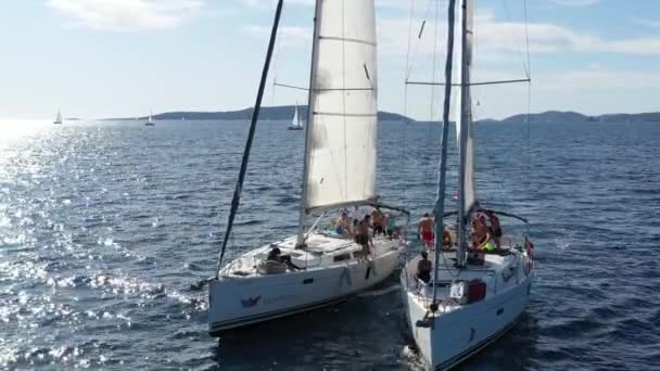 克罗地亚，亚得里亚海，2019年9月18日：两艘帆船彼此距离很近，年轻人在游艇上玩得很开心，年轻人挂在船与船之间的绳索上，岛上有背风，水面上有阳光反射 — 图库视频影像