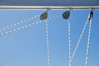 Yelkenli halatlarının güneşli havalarda yakın görüntüsü, direkte makaralar ve ipler, yat sporu, gemi ekipmanları, arka planda deniz var.