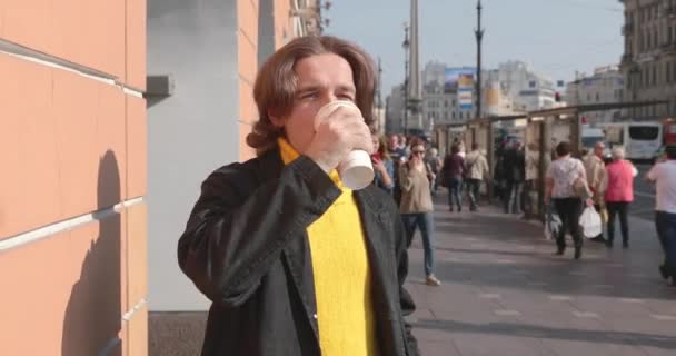 Los hombres guapos está esperando a alguien, bebe café, se vistió con un suéter amarillo y impermeable negro, la calle de San Petersburgo y un montón de peatones están en el fondo, día soleado — Vídeo de stock