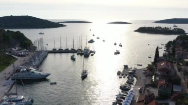 Hırvatistan, güneş batımında Solta adasındaki marina manzarası, bir sürü yelkenli, turuncu renkli çatılar, suya yansıyan güneş, yeşil ağaçlar.
