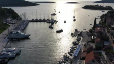 Hırvatistan, güneş batımında Solta adasındaki marina manzarası, bir sürü yelkenli, turuncu renkli çatılar, suya yansıyan güneş, yeşil ağaçlar.