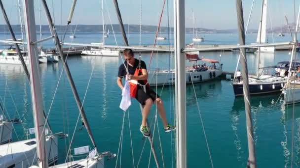 Κροατία, μαρίνα Καστέλα, 15 Σεπτεμβρίου 2019: Σημείο προβολής του ναύτη που κρέμεται σε μια κούνια σε ένα κατάρτι και δένει σημαίες, συμμετέχων σε ιστιοπλοϊκή ρεγκάτα, ο κόσμος περιμένει τον επερχόμενο αγώνα — Αρχείο Βίντεο