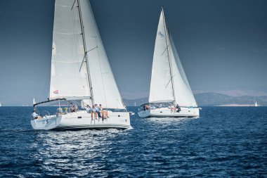 Yelkenli yarışı, yelkenli yelkenli yarışı, sudaki yelkenli yansıması, yoğun rekabet, arka teknelerde tekne sayısı, parlak renkler, arka planda ada