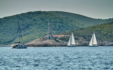 Yelkenli yarışı, yelkenli yelkenli yarışı, sudaki yelkenli yansıması, yoğun rekabet, arka teknelerde tekne sayısı, parlak renkler, arka planda ada