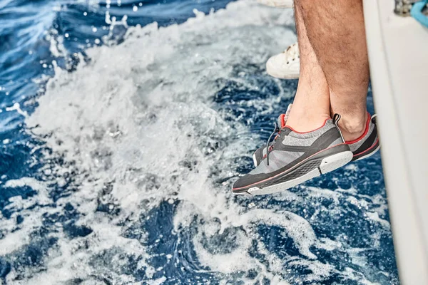 Los pies del marinero vestido con calzado de yate cuelgan de una tabla de barco sobre el agua azul, salpicaduras, manchas de luz en el agua, vela, tiempo soleado, reflejos del sol, vacaciones —  Fotos de Stock
