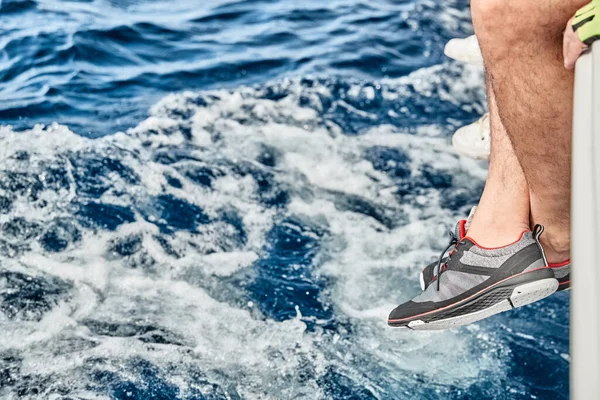 Los pies del marinero vestido con calzado de yate cuelgan de una tabla de barco sobre el agua azul, salpicaduras, manchas de luz en el agua, vela, tiempo soleado, reflejos del sol, vacaciones —  Fotos de Stock