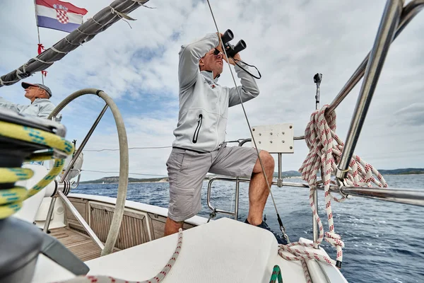 Croacia, Mar Adriático, 19 de septiembre de 2019: El brutal capitán del barco está en un volante, mira en el vidrio de campo, chaqueta de color azul, velero competir en una regata de vela, tiempo nublado — Foto de Stock