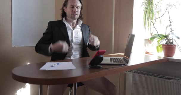 De mooie zakenman in een zakenpak en zonder broek blijft thuis aan een keukentafel, hij werkt op de laptop, houdt conferentie, verre werk tijdens een pandemie Covid-19, quarantaine — Stockvideo