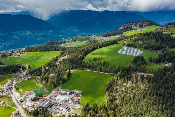 Вид с воздуха на ферму солнечных батарей на зеленых склонах гор Италии, Трентино, огромные облака над долиной, крыши домов поселений, зеленые луга, чистая энергия, энергия солнца — стоковое фото