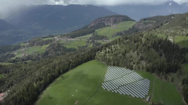 Widok z powietrza na farmę baterii słonecznych na zielonych zboczach gór Włoch, Trentino, ogromne chmury nad doliną, dachy domów osiedli, zielone łąki, czysta energia, energia słońca — Wideo stockowe