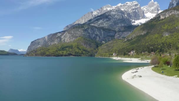 Det osannolika luftlandskapet i byn Molveno, Italien, drönare flyger lågt över azurvatten i sjön, en tom strand, snötäckta sluttningar av berg på bakgrunden, soligt väder, kust — Stockvideo