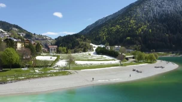 A valószínűtlen légi táj falu Molveno, Olaszország, drón repül alacsony azúrvíz a tó, egy üres strand, havas lejtők a hegy a háttérben, napos időjárás, tengerpart — Stock videók