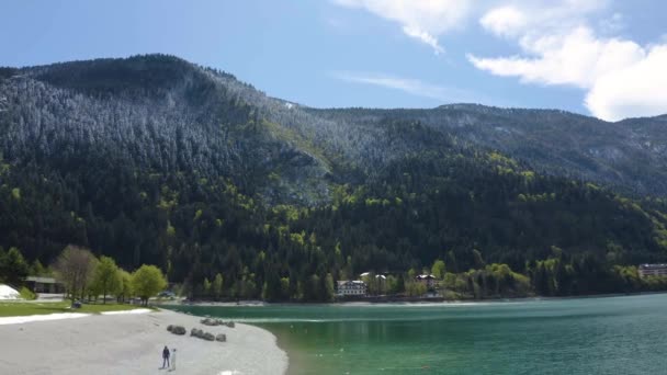 Die unwahrscheinliche Luftlandschaft des Dorfes Molveno, Italien, Drohne fliegt tief über das azurblaue Wasser des Sees, ein leerer Strand, schneebedeckte Fichten an Berghängen im Hintergrund, sonniges Wetter, Küste — Stockvideo