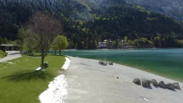 A valószínűtlen légi táj falu Molveno, Olaszország, drón repül alacsony azúrvíz tó, egy üres strand, havas lucfenyő lejtőin a hegy a háttérben, napos időjárás, tengerpart — Stock videók