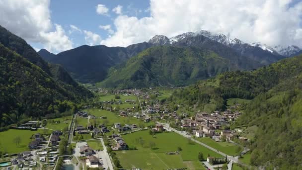 A paisagem aérea improvável de aldeia Molveno, Itália, água azul de lago, praia vazia, montanhas cobertas de neve Dolomites no fundo, topo de telhado de chalé, tempo ensolarado, um cais, litoral , — Vídeo de Stock