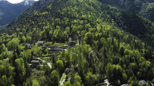 Házak és különböző épületek állnak a lejtőn a hegy borított zöld fák, lucfenyő a napsütéses időjárás, drón repül körül, a hegyek Olaszország, Trentino, Molveno — Stock videók
