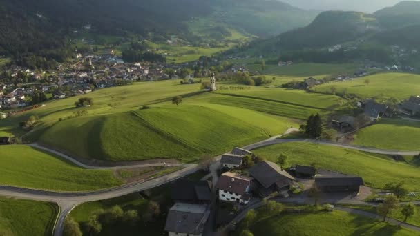 Pandangan udara dari lembah indah dengan kapel di Bolzano saat matahari terbenam, Trentino, padang rumput hijau yang menakjubkan dari pegunungan Italia, atap rumah, Dolomites di latar belakang, drone terbang di sekitar kapel — Stok Video