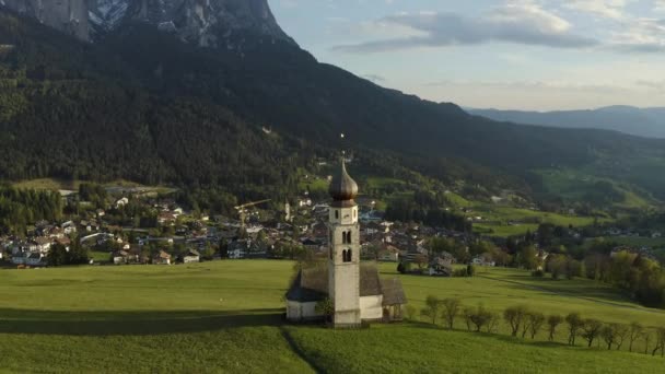 Flygfoto över pittoresk dal med kapell i Bolzano vid solnedgången, Trentino, fantastiska gröna ängar i bergen i Italien, tak toppar av hus, Dolomiter på bakgrunden, drönare flyger runt kapell — Stockvideo