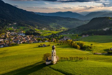 Gün batımında Bolzano 'da şapeli olan pitoresk vadinin havadan görünüşü, Trentino, İtalya dağlarının muhteşem yeşil çayırları, evlerin çatıları, arka planda Dolomitler, şapelin etrafında insansız hava aracı uçuşları.