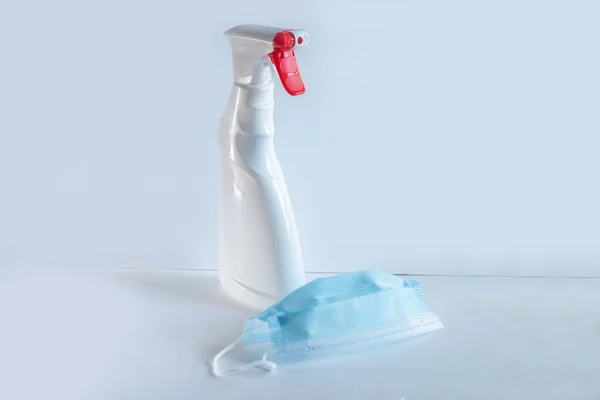 Lo spruzzo di bottiglia di colore bianco con il pulsante rosso e la maschera medica di colore azzurro è su un tavolo del colore bianco, una serie durante una pandemia per disinfezione, protezione di sistema respiratorio — Foto Stock