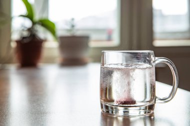 Bardağa atılan köpüklü tablet şeffaf bir bardağın içinde, cam masanın üzerinde, soğuk kırmızı şarap için bir hap, kabarcıklar yükseliyor, bardaktan fışkırıyor, çiçekler arka planda, köpükler...