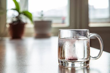 Bardağa atılan köpüklü tablet şeffaf bir bardağın içinde, cam masanın üzerinde, soğuk kırmızı şarap için bir hap, kabarcıklar yükseliyor, bardaktan fışkırıyor, çiçekler arka planda, köpükler...