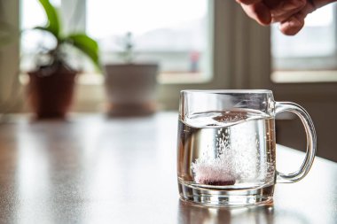 Bardağa atılan köpüklü tablet şeffaf bir bardağın içinde, cam masanın üzerinde, soğuk kırmızı şarap için bir hap, kabarcıklar yükseliyor, bardaktan fışkırıyor, çiçekler arka planda, köpükler...