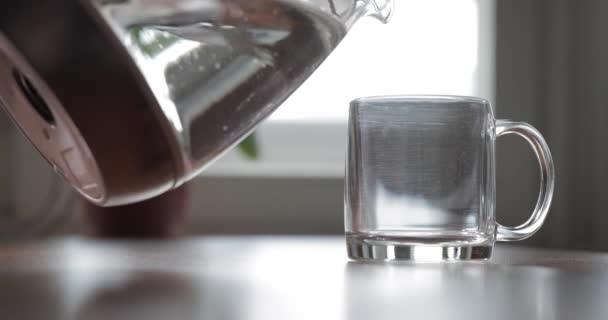 El cristal transparente de cristal está sobre una mesa, el agua se vierte de una tetera en un vaso, las flores están en el fondo — Vídeos de Stock