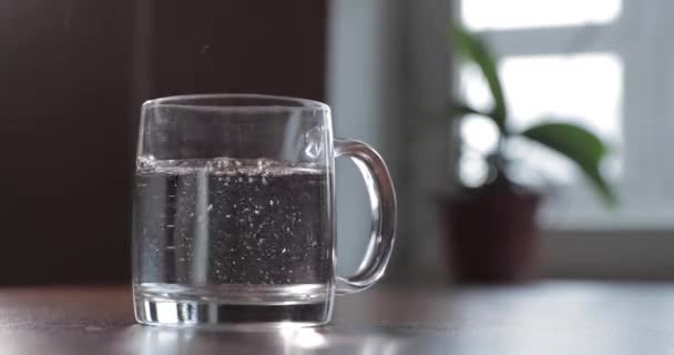 Brillante tableta tirar en un vidrio transparente de vidrio, el vidrio está en una mesa, una píldora para el frío de color clarete, burbujas se levantan, salpicaduras despegan de un vidrio, flores está en el fondo, espuma — Vídeos de Stock