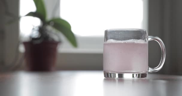 Sprankelende tablet gooien in een glas transparant glas, het glas is op een tafel, een pil voor de kou van Claret kleur, bellen opstaan, spatten opstijgen van een glas, bloemen is op de achtergrond, schuim — Stockvideo