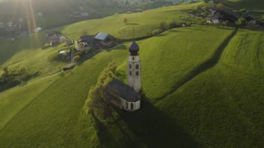 Gün batımında Bolzano 'da şapeli olan pitoresk vadinin havadan görünüşü, Trentino, İtalya dağlarının muhteşem yeşil çayırları, evlerin çatıları, arka planda Dolomitler, şapelin etrafında insansız hava aracı uçuşları.