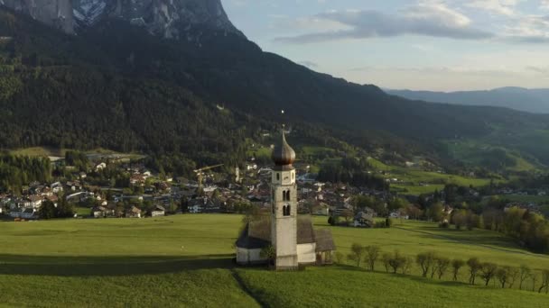 Vista aérea del pintoresco valle con capilla en Bolzano al atardecer, Trentino, increíbles prados verdes de las montañas de Italia, azoteas de casas, Dolomitas en el fondo, drones vuela alrededor de la capilla — Vídeos de Stock