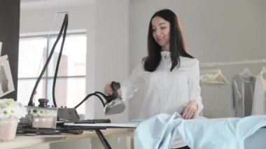 Genç kızın yavaş videosu ütülüyor. Elbiseleri askıya asıyor, dumanlar dikey durumda, buharlı profesyonel bir ütü, dikiş üretimi, dikiş stüdyosu, ajans...