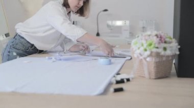 Moda tasarımcısının yavaş çekim videosu kıvrımlı ve desenli bir masada çalışıyor, kumaşı kesiyor, etrafında makas, santimetre, dikiş makinesi ve arka planda kukla, pürüzsüz bir hareket var.
