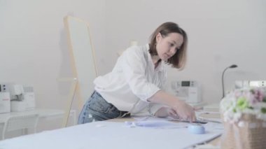 Moda tasarımcısının yavaş çekim videosu kıvrımlı ve desenli bir masada çalışıyor, kumaşı kesiyor, etrafında makas, santimetre, dikiş makinesi ve arka planda kukla, pürüzsüz bir hareket var.
