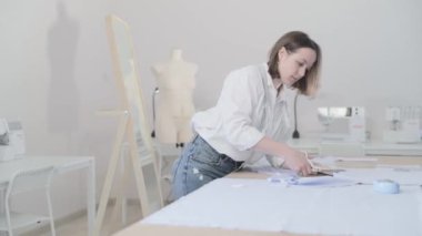 Moda tasarımcısının yavaş çekim videosu kıvrımlı ve desenli bir masada çalışıyor, kumaşı kesiyor, etrafında makas, santimetre, dikiş makinesi ve arka planda kukla, pürüzsüz bir hareket var.
