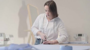 Moda tasarımcısının yavaş çekim videosu kıvrımlı ve desenli bir masada çalışıyor, kumaşı kesiyor, etrafında makas, santimetre, dikiş makinesi ve arka planda kukla, pürüzsüz bir hareket var.