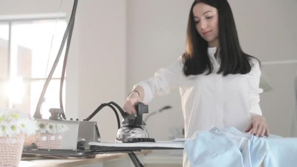 Αργό βίντεο με το νεαρό όμορφο κορίτσι σιδερώνει τα ρούχα που κρέμονται σε μια κρεμάστρα παλτό στο κιγκλίδωμα, ατμούς σε κάθετη κατάσταση, ένα επαγγελματικό σίδερο με ατμό, ράψιμο παραγωγής, ράψτε στούντιο, πρακτορείο — Αρχείο Βίντεο