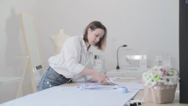 Slow motion video of the fashion designer works at a table with curves and a pattern, she are cutting fabric, around lies scissors, centimeter, sew machine and dummy on the background, Smooth movement — Stock Video