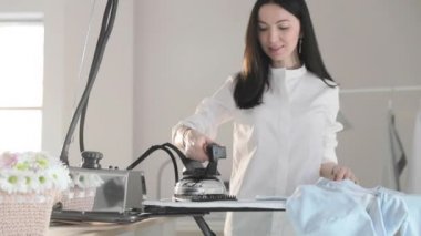 Genç kızın yavaş videosu ütülüyor. Elbiseleri askıya asıyor, dumanlar dikey durumda, buharlı profesyonel bir ütü, dikiş üretimi, dikiş stüdyosu, ajans...
